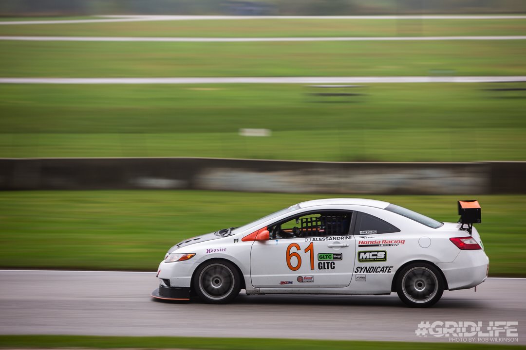 A white 9livesracing Civic Coupe Wang kit '06-11, with the number 61 displayed prominently on the side, speeds along a wet racetrack, featuring extruded aluminum wings for an optimal lift-to-drag ratio. The car showcases various sponsor logos and netted windows. In the blurred grassy background, "#GRIDLIFE" is visible in the bottom right corner.