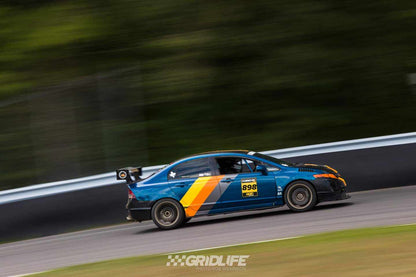 A blue and orange Civic Sedan Wang kit '06-11 FA5 by 9livesracing, marked with the number 898, speeds along a track during a "Gridlife" event. The background is blurred, emphasizing the car's motion. Trees and a barrier wall are visible in the distance, while airfoils on the car optimize its lift-to-drag ratio for maximum performance.