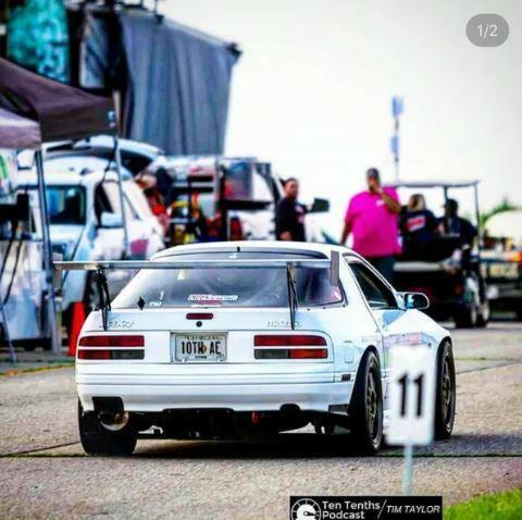 An RX7 Big Wang Kit '85-92 FC by 9livesracing, featuring a large aluminum wing, is parked on a race track. The white performance racing car has a license plate reading "1OTHSAC" and is adorned with various stickers on the rear window. In the background, several people and vehicles are visible near a tent setup.