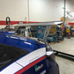 A blue sports car featuring a striking 9livesracing Civic Type-R Wang kit 2017+ FK8 rear wing spoiler is parked in a garage. The interior of the garage is filled with various tools, equipment, and a workspace with shelves and boxes in the background. The car has decals and logos on the side, showcasing modifications and sponsors aimed at optimizing down-force.