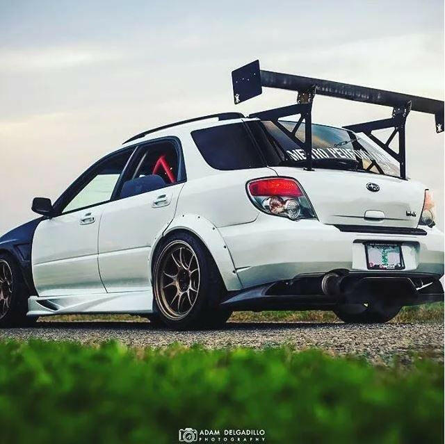 A white modified hatchback car is parked on a paved road beside green grass. The Subaru Impreza Wagon, equipped with the 9livesracing Impreza Wagon Big Wang kit '00-07 GG, boasts a large rear wing, gold rims, adjustable downforce via performance airfoils, a rear diffuser, and an interior roll cage. The sky in the background is clear with a hint of sunset.