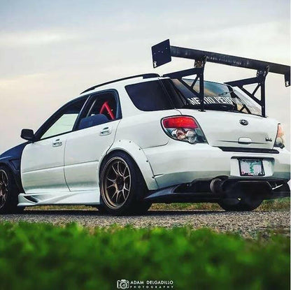 A white modified hatchback car is parked on a paved road beside green grass. The Subaru Impreza Wagon, equipped with the 9livesracing Impreza Wagon Big Wang kit '00-07 GG, boasts a large rear wing, gold rims, adjustable downforce via performance airfoils, a rear diffuser, and an interior roll cage. The sky in the background is clear with a hint of sunset.