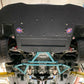 View from underneath a 370z Rocket Nose v2 Splitter ’08-20 Z34, by 9livesracing, showing the rear undercarriage. Visible are large tires, suspension components, exhaust system, and a diffuser with performance stickers enhancing downforce. The garage ceiling and hydraulic lift arms can also be seen.