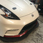 Close-up image of the front corner of a white Nissan 370z featuring a sleek headlight design, a black front lip with red accents, and a Nissan emblem on the grille. The 9livesracing 370z Rocket Nose v2 Splitter ’08-20 Z34 enhances downforce for superior performance. The car is parked in what appears to be a garage or workshop with tools visible in the background.