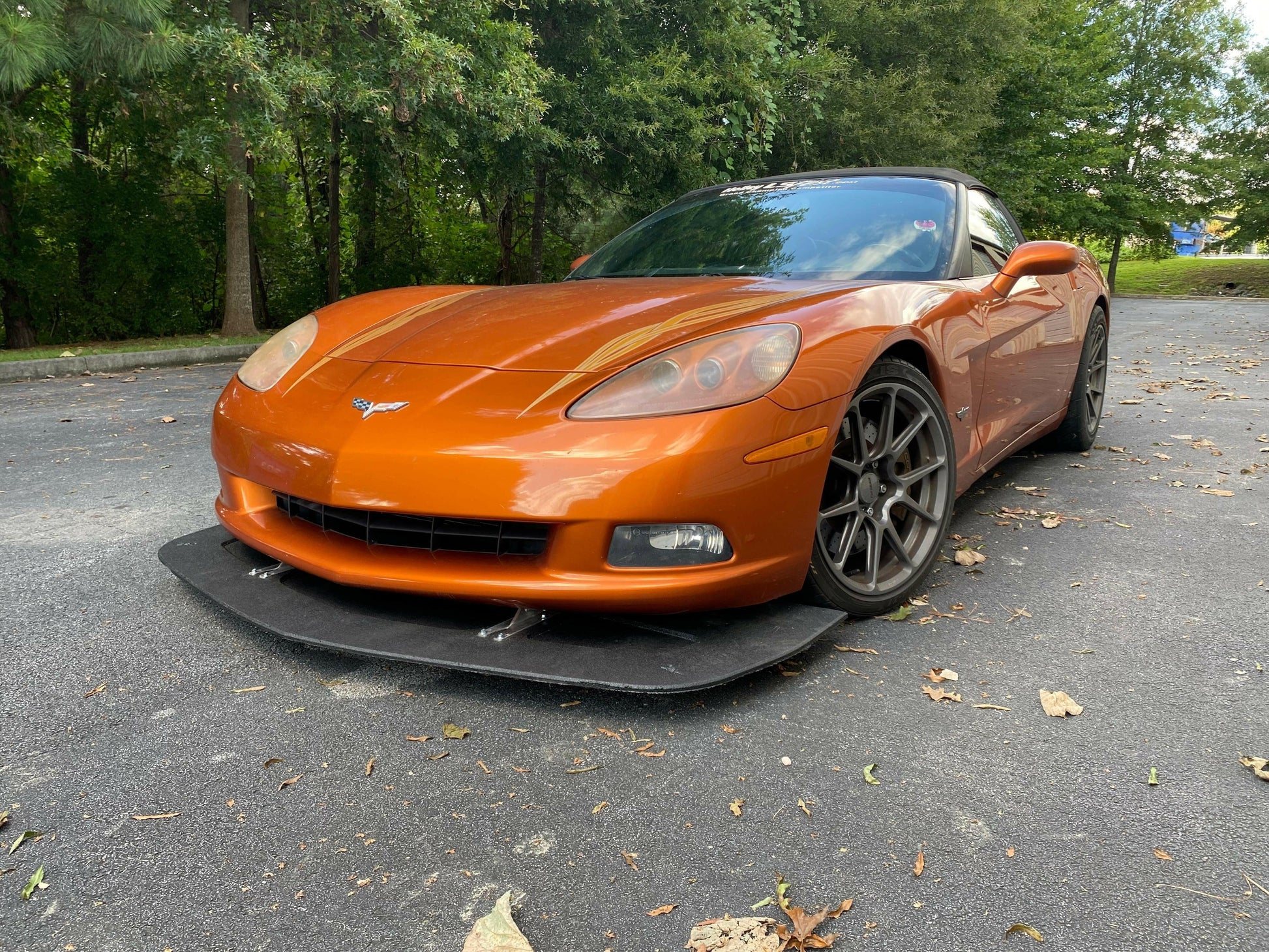 A sleek orange sports car, equipped with a Corvette Splitter - C6 & C5 ‘05-13 by 9livesracing, is parked on a pavement scattered with dry leaves. The background features a dense cluster of trees and greenery. The car's low profile and aerodynamic design, accentuated by its carbon components, are highlighted in the image.