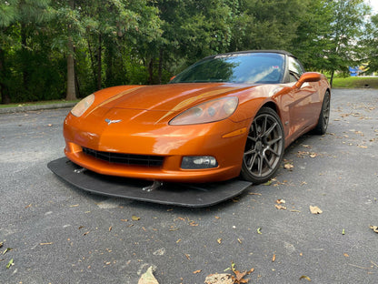 A sleek orange sports car, equipped with a Corvette Splitter - C6 & C5 ‘05-13 by 9livesracing, is parked on a pavement scattered with dry leaves. The background features a dense cluster of trees and greenery. The car's low profile and aerodynamic design, accentuated by its carbon components, are highlighted in the image.