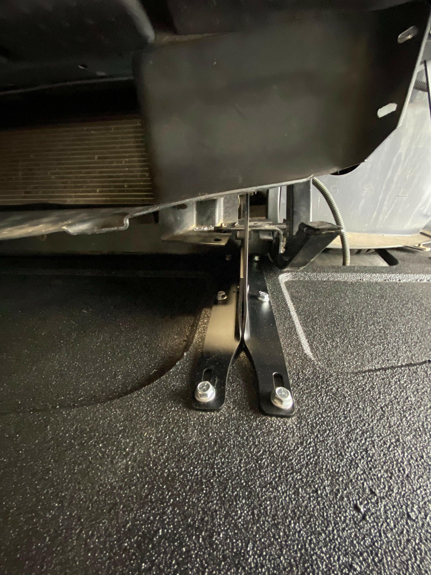 Close-up view of the 9livesracing Corvette Splitter - C6 & C5 ‘05-13 securing a vehicle’s component. The splitter, crafted with CNC cut pockets, is attached with bolts and situated beneath a larger black assembly, with part of a radiator visible in the background. The surface below the splitter is textured and black.