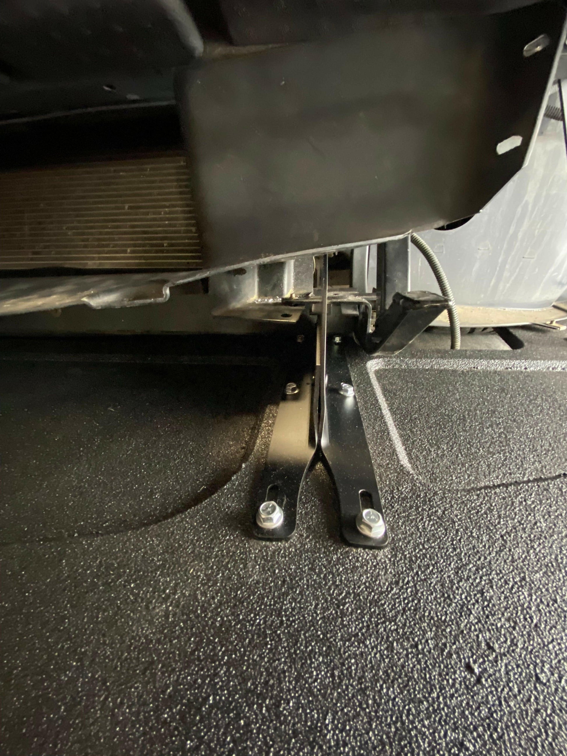 Close-up view of the 9livesracing Corvette Splitter - C6 & C5 ‘05-13 securing a vehicle’s component. The splitter, crafted with CNC cut pockets, is attached with bolts and situated beneath a larger black assembly, with part of a radiator visible in the background. The surface below the splitter is textured and black.