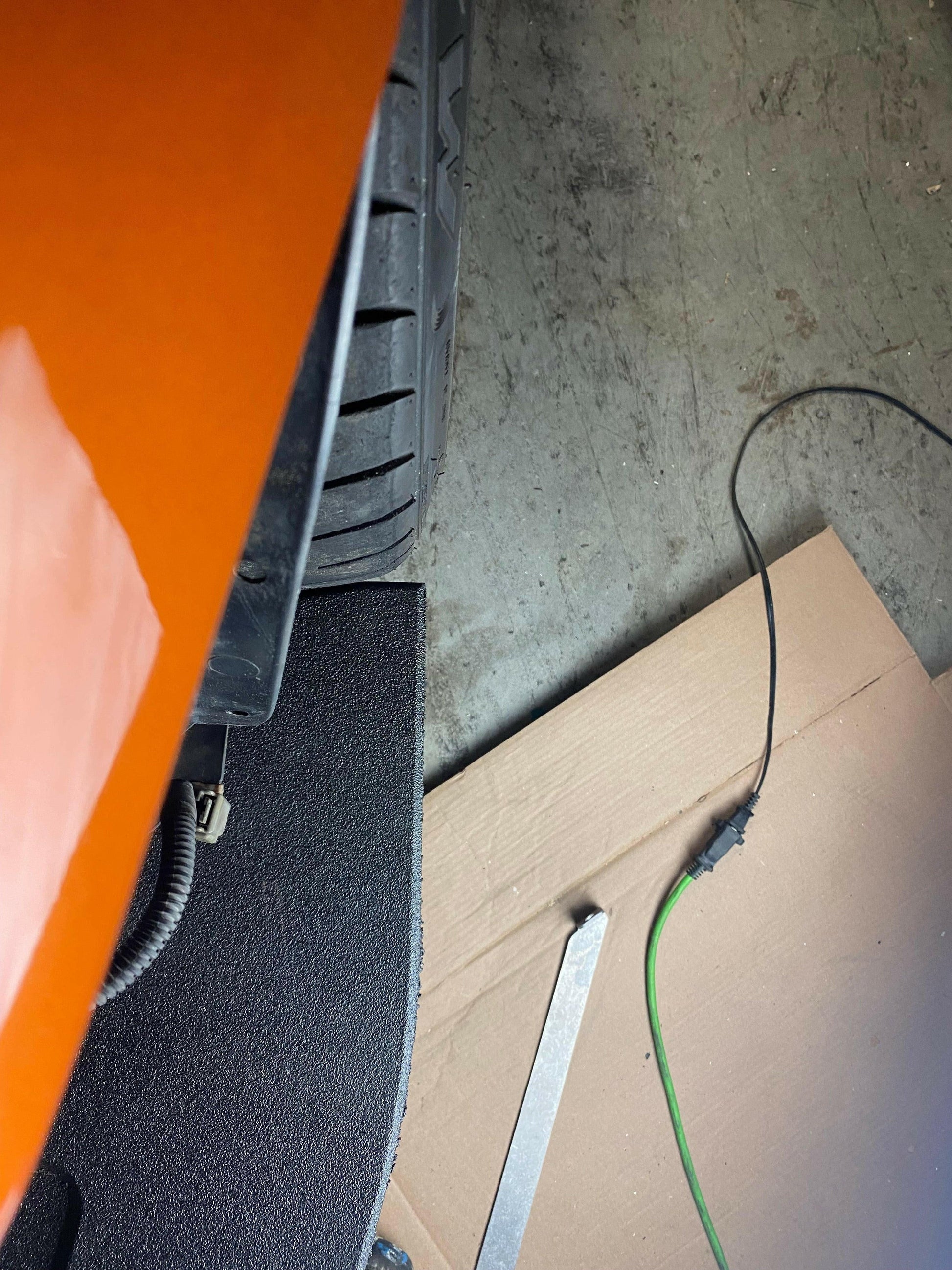 Close-up view of the rear part of an orange car, focusing on the tire, part of the wheel well, and adjacent ground. Near the tire are a piece of cardboard, a metal bar with precise CNC cut pockets from 9livesracing's Corvette Splitter - C6 & C5 ‘05-13 collection, and a green cable with black connectors lying on a concrete surface.