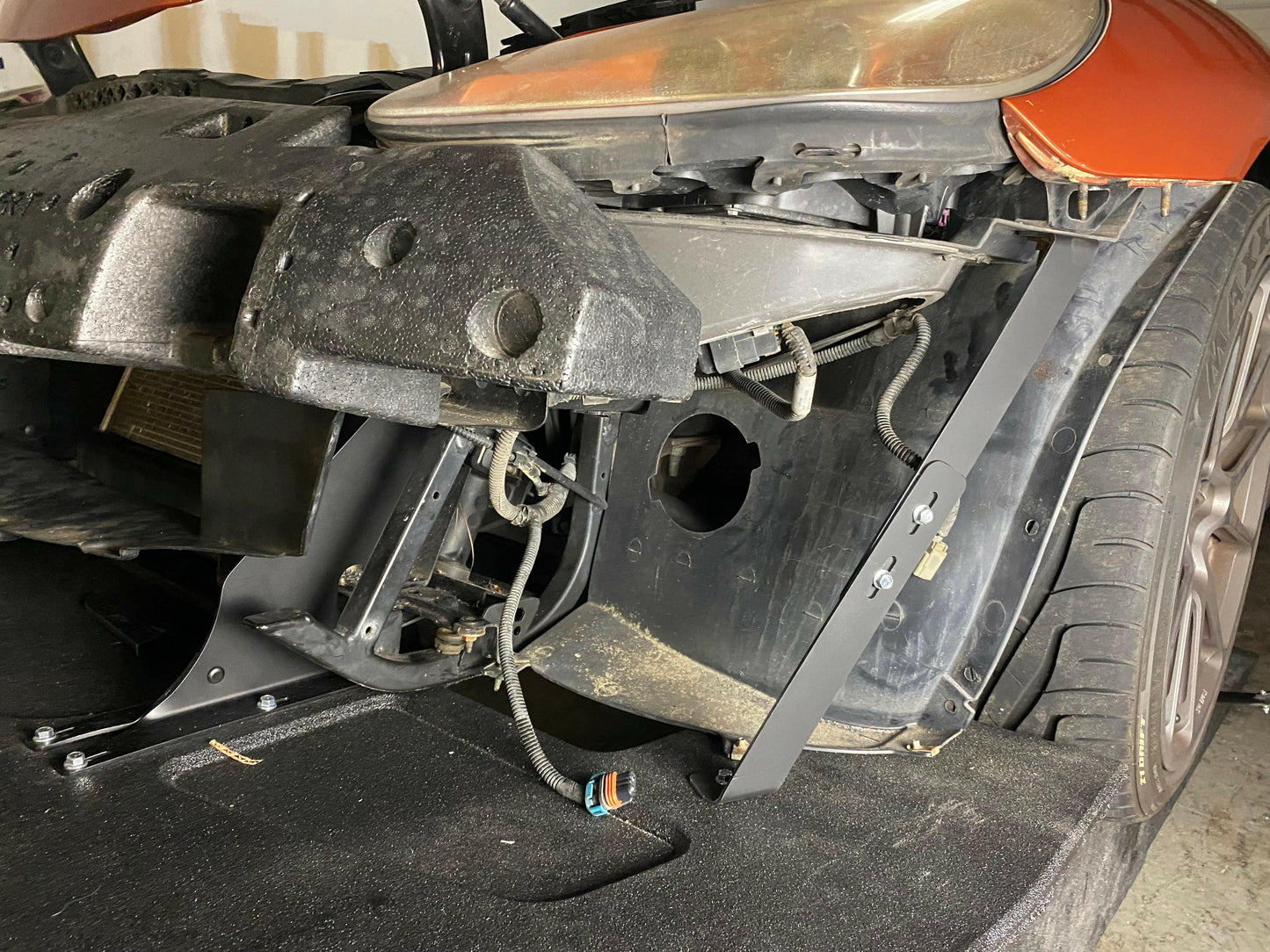 A Corvette with its front bumper removed, exposing internal components and wiring. The headlight area is visible, and the car's wheel remains attached. The image shows various mechanical parts, including low friction polymer brackets and CNC cut pockets amid wires, against the backdrop of a garage. This particular model features the 9livesracing Corvette Splitter - C6 & C5 ‘05-13.