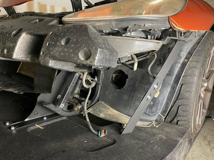A Corvette with its front bumper removed, exposing internal components and wiring. The headlight area is visible, and the car's wheel remains attached. The image shows various mechanical parts, including low friction polymer brackets and CNC cut pockets amid wires, against the backdrop of a garage. This particular model features the 9livesracing Corvette Splitter - C6 & C5 ‘05-13.