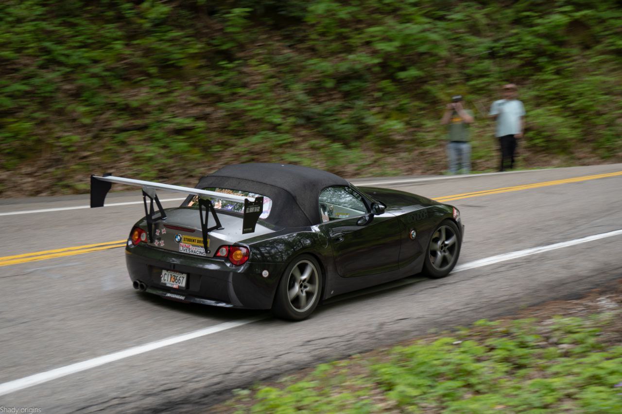 A black BMW Z4 convertible sports car, equipped with a 9livesracing Big Wang Kit from the 2003-08 (E85) series, speeds down a forested road. Two people stand on the roadside, one capturing a photo. The rear of the car is adorned with various stickers from Nine Lives Racing, and an "ADO 0894" license plate is visible.