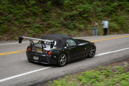 A black BMW Z4 convertible sports car, equipped with a 9livesracing Big Wang Kit from the 2003-08 (E85) series, speeds down a forested road. Two people stand on the roadside, one capturing a photo. The rear of the car is adorned with various stickers from Nine Lives Racing, and an "ADO 0894" license plate is visible.