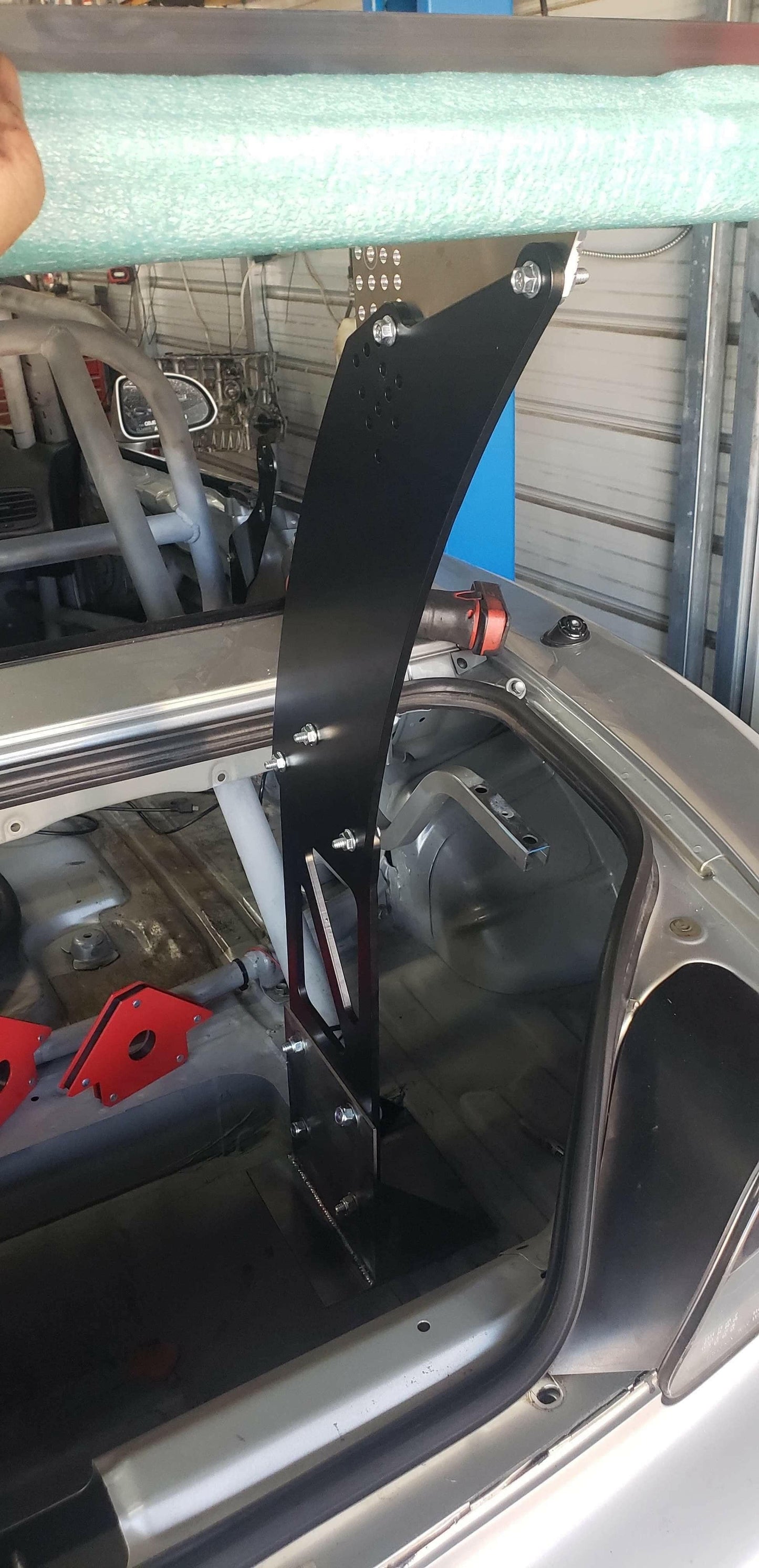 A black metal bracket from the S2000 Big Wang Chassis Mount Kit '99-09 AP1/AP2 by 9livesracing is installed inside a Honda S2000 with a roll cage. This bracket is securely bolted to the frame and extends above the car's rear window. A green cylindrical object, partially visible above the bracket and held by a person's hand, is also present.

