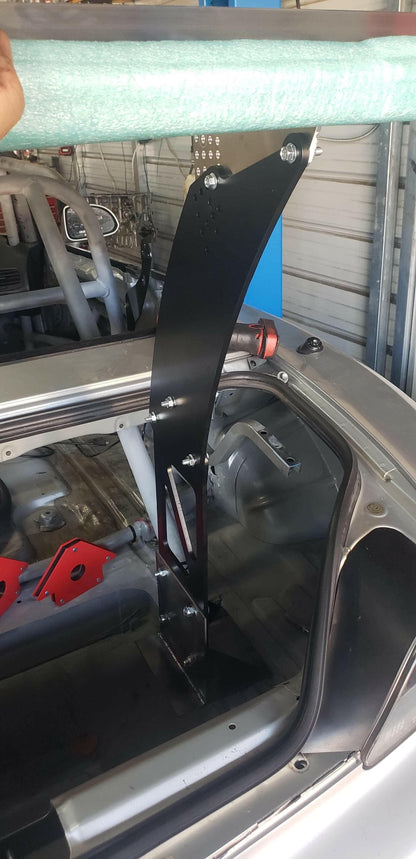A black metal bracket from the S2000 Big Wang Chassis Mount Kit '99-09 AP1/AP2 by 9livesracing is installed inside a Honda S2000 with a roll cage. This bracket is securely bolted to the frame and extends above the car's rear window. A green cylindrical object, partially visible above the bracket and held by a person's hand, is also present.

