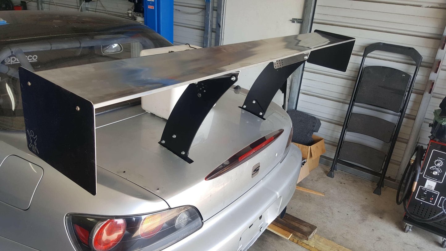 A silver Honda S2000, equipped with the 9livesracing S2000 Big Wang Chassis Mount Kit '99-09 AP1/AP2 featuring a large black and silver rear wing spoiler, is parked in a garage. In the background, tools and equipment are visible, including a folding chair, a cart with various tools, and an empty cardboard box. This setup optimizes down-force for improved handling.