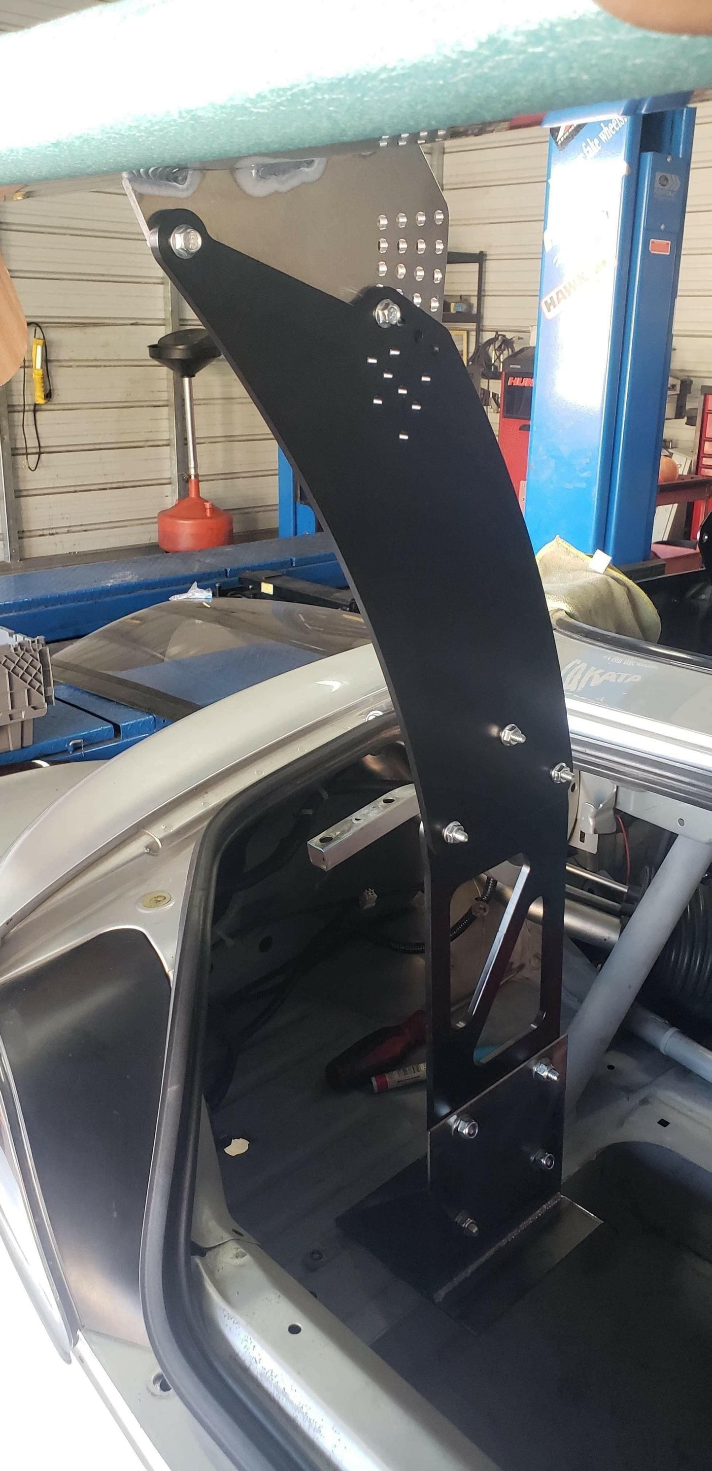 Close-up of a Honda S2000 interior showcasing the 9livesracing S2000 Big Wang Chassis Mount Kit '99-09 AP1/AP2. The large, black metal mount is securely bolted to the frame and extends vertically from the floor behind the driver's seat to the ceiling. It features multiple bolt attachments and has a perforated design at the top. The background reveals a workshop setting.