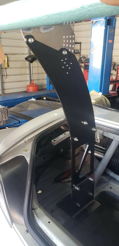 Close-up of a Honda S2000 interior showcasing the 9livesracing S2000 Big Wang Chassis Mount Kit '99-09 AP1/AP2. The large, black metal mount is securely bolted to the frame and extends vertically from the floor behind the driver's seat to the ceiling. It features multiple bolt attachments and has a perforated design at the top. The background reveals a workshop setting.