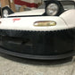 A white Mazda Miata with pop-up headlights and a large black front splitter from 9livesracing stands parked in a garage. The car, featuring a red emblem on the front, utilizes its Rocket Nose design for enhanced downforce. Various items are scattered in the background while the photo highlights the front bumper and splitter, which is designed for Mazda Miatas from 1990-05 NA/NB models.