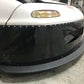 Close-up of the front end of a car featuring a white hood, yellow-tinted headlights, and a black front bumper with rivets and a textured gray Mazda Miata Splitter (1990-05 NA/NB) by 9livesracing. The car's aggressive Rocket Nose design enhances downforce, parked on a concrete floor in a garage or workshop with blurred tools and equipment in the background.