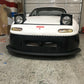 A white Mazda Miata, equipped with raised pop-up headlights and a sleek rocket nose, is parked in a garage. The car features a black front bumper and proudly displays a red emblem on the hood. Assorted items and equipment are visible in the background, enhancing its high-performance aura.
