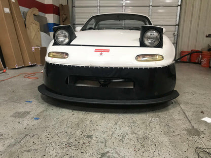A white Mazda Miata, equipped with raised pop-up headlights and a sleek rocket nose, is parked in a garage. The car features a black front bumper and proudly displays a red emblem on the hood. Assorted items and equipment are visible in the background, enhancing its high-performance aura.