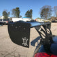 A black sports car, equipped with a large 9livesracing carbon wing, sits in a parking lot. The spoiler, designed to maximize down-force, prominently features the distinctive 9livesracing logo of a figure with spread arms. In the background, other parked vehicles, buildings, and tall trees stand under a clear blue sky.