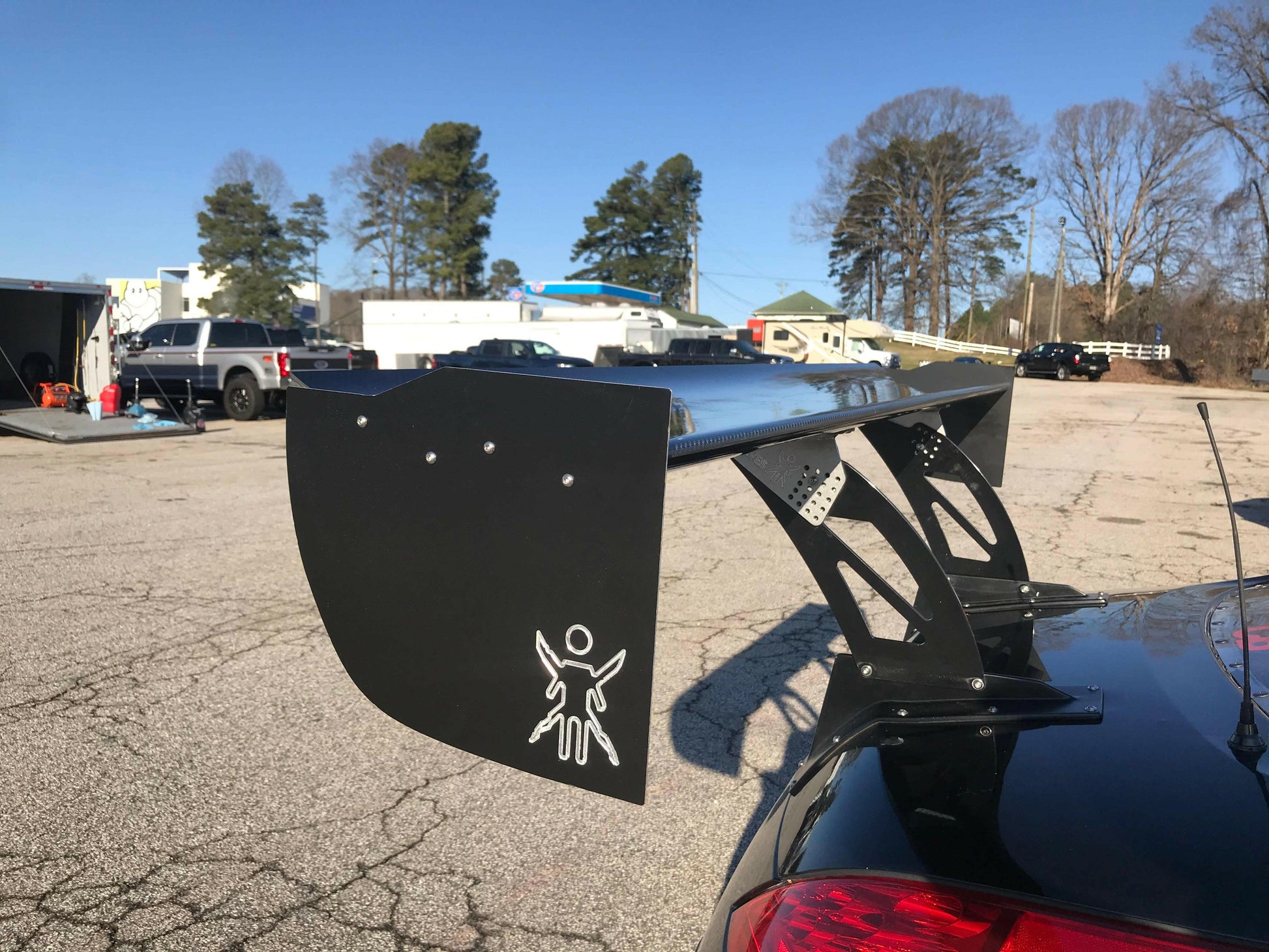 A black sports car, equipped with a large 9livesracing carbon wing, sits in a parking lot. The spoiler, designed to maximize down-force, prominently features the distinctive 9livesracing logo of a figure with spread arms. In the background, other parked vehicles, buildings, and tall trees stand under a clear blue sky.