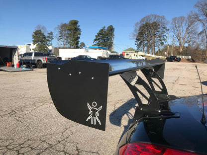 A black sports car, equipped with a large 9livesracing carbon wing, sits in a parking lot. The spoiler, designed to maximize down-force, prominently features the distinctive 9livesracing logo of a figure with spread arms. In the background, other parked vehicles, buildings, and tall trees stand under a clear blue sky.