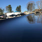 A close-up shot of a sleek, reflective surface, likely a car hood featuring a 9livesracing carbon wing. Trees and parked vehicles are visible in the background under a clear blue sky, capturing a crisp day with bare trees and several black vehicles lined up.