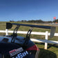A rear view of a black sports car with a large 9livesracing carbon wing, parked near a white fence. The logo "9livesracing" is visible on the car. A grassy area and a racetrack with barriers are in the background under a clear blue sky.