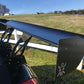 Close-up of a large, black pre-preg 9livesracing carbon wing on a car, set against a rural backdrop with a green field and clear blue sky. The spoiler features the distinct 9livesracing logo with a stylized figure on one side panel. A white wooden fence runs parallel to the car in the background.