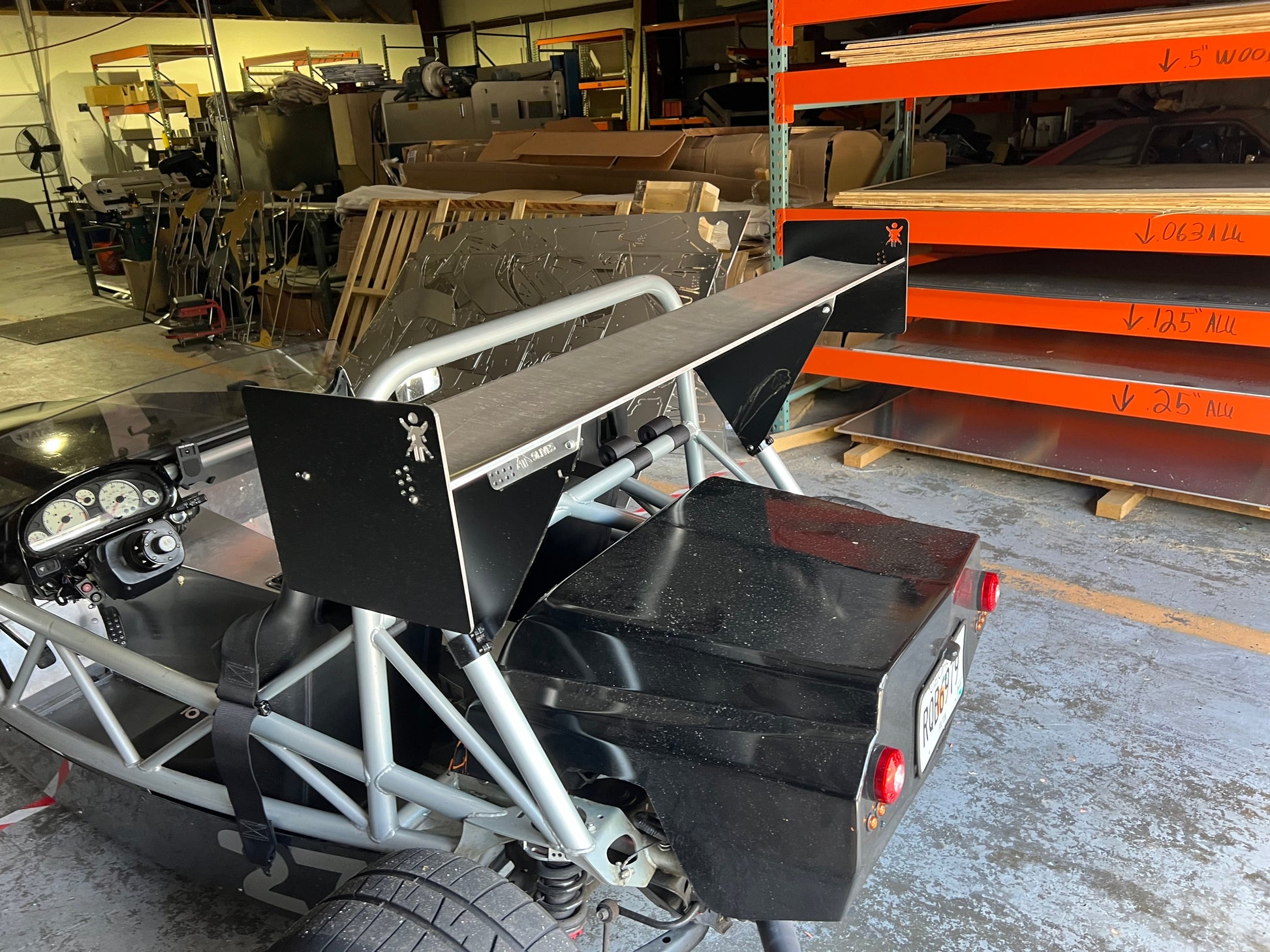 An Exocet v2 Big Wang Kit, a minimalist and custom-built open-frame car by 9livesracing, featuring a striking rear wing, is pictured in a workshop. Surrounding the vehicle are various automotive tools, stacked shelves, and metallic panels. The car’s interior detailing and rear lights are visible, suggesting its GT3 performance capabilities.
