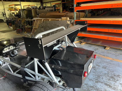 An Exocet v2 Big Wang Kit, a minimalist and custom-built open-frame car by 9livesracing, featuring a striking rear wing, is pictured in a workshop. Surrounding the vehicle are various automotive tools, stacked shelves, and metallic panels. The car’s interior detailing and rear lights are visible, suggesting its GT3 performance capabilities.
