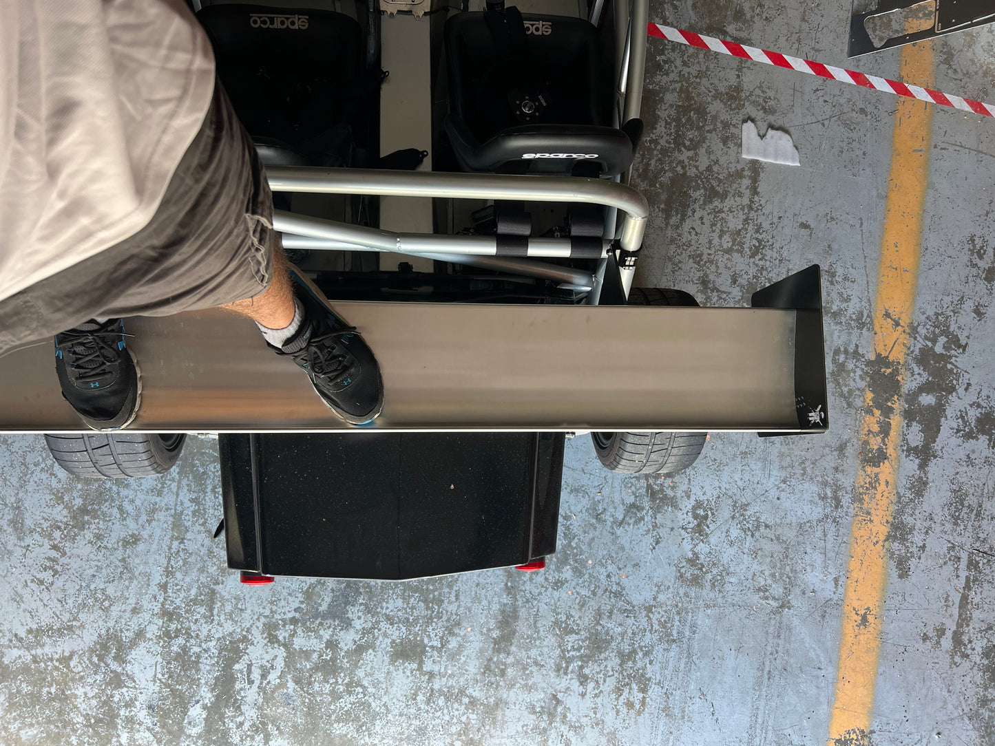 A person stands on the rear wing of an Exocet v2 Big Wang Kit by 9livesracing sports car, with the view looking down from above. The person is wearing shorts and black shoes. The car's rear wheels, seats, and wing are visible, along with a section of the ground marked with yellow and red lines.