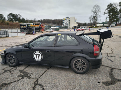 A black race car is parked in a cracked lot under an overcast sky, proudly displaying the number 17 on its door. It features a Civic Hatch EK Wang kit from 9livesracing, complete with a Big Wang rear wing and modified body parts for enhanced down-force. Trees, trailers, and buildings are visible in the background.