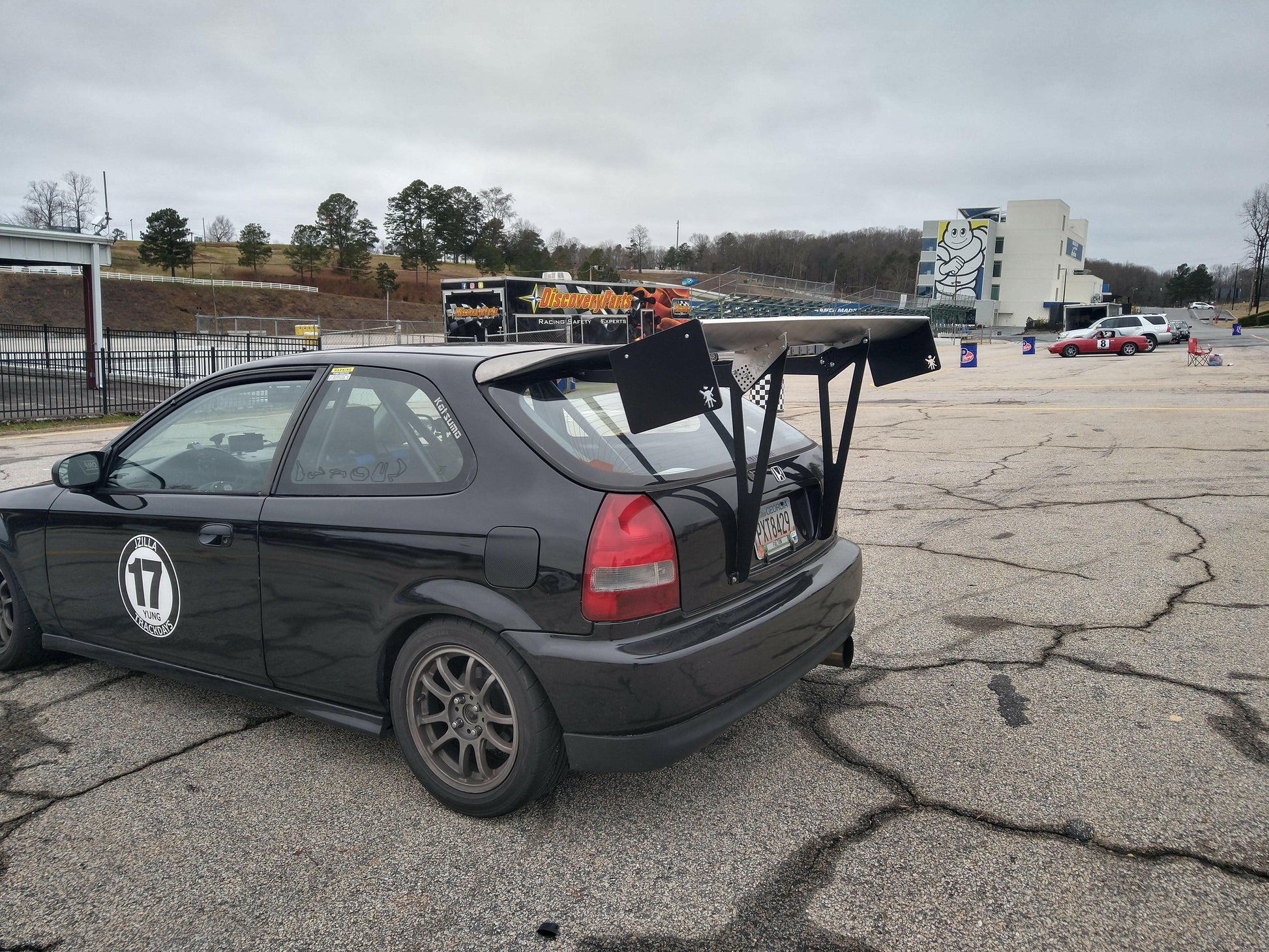 A 9livesracing Civic Hatch Ek Wang kit 96-2000 with the number 17 on its side is parked in a cracked asphalt area. The car features a large, aluminum wing for maximum down-force and dark alloy wheels. In the background, various vehicles and a partially visible grandstand can be seen on a cloudy day.