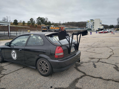 A 9livesracing Civic Hatch Ek Wang kit 96-2000 with the number 17 on its side is parked in a cracked asphalt area. The car features a large, aluminum wing for maximum down-force and dark alloy wheels. In the background, various vehicles and a partially visible grandstand can be seen on a cloudy day.