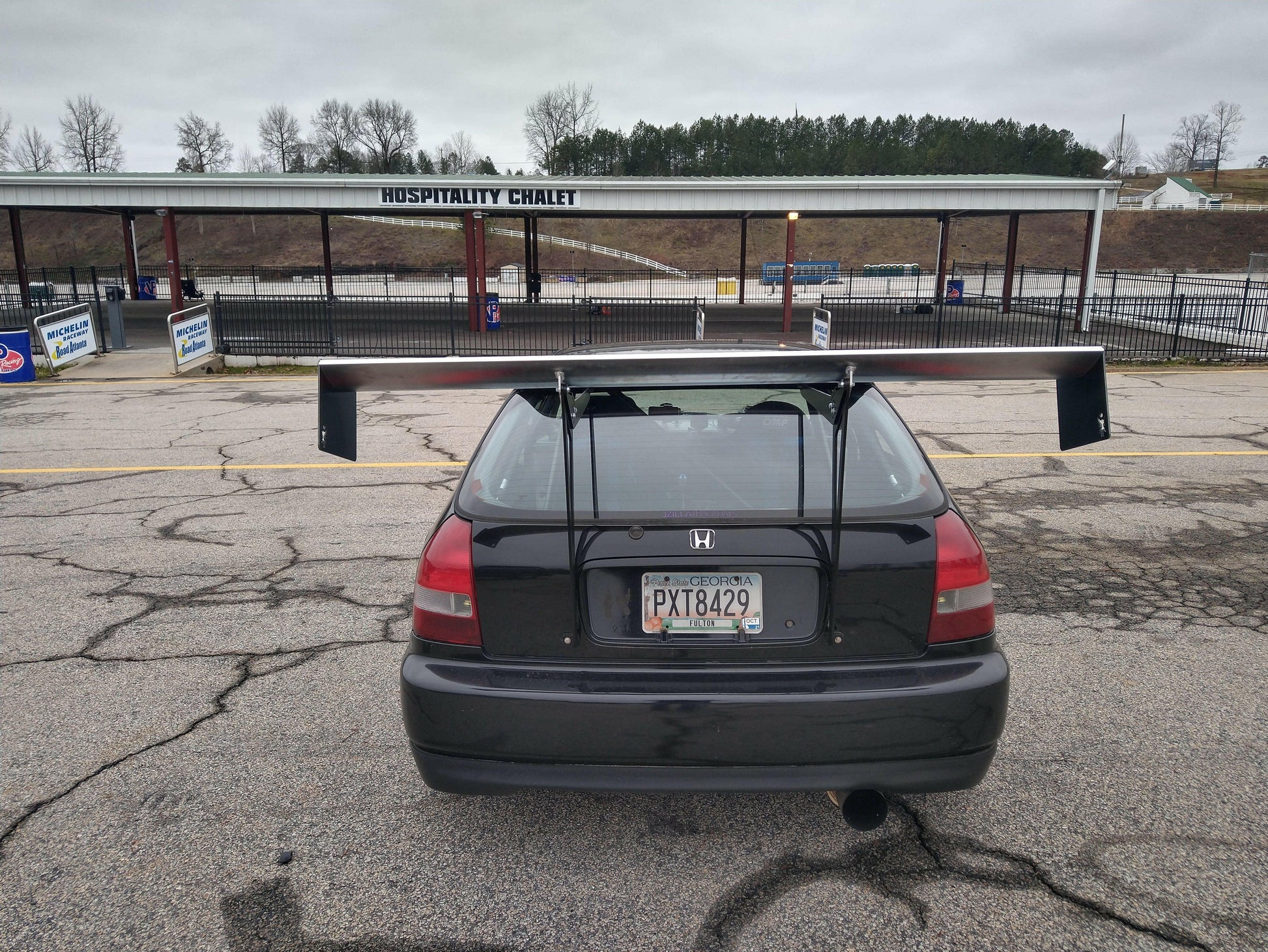 A black Honda Civic Hatch Ek featuring a large spoiler from the 9livesracing Wang kit (96-2000) is parked in front of a structure labeled "Hospitality Chalet." The overcast sky and distant trees cast a gray tone over the image. The car bears a Georgia license plate with the number PXT8429.