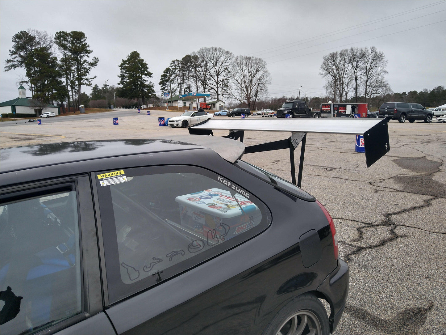 A car outfitted with the Civic Hatch EK Wing Kit 96-2000 from 9livesracing is parked in a lot. Visible through the window, a box containing a racing console sits in the backseat. Other vehicles and trees are visible in the background on this cloudy day.