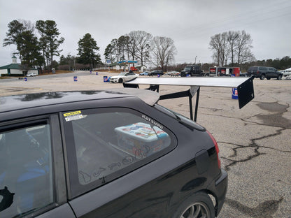 A car outfitted with the Civic Hatch EK Wing Kit 96-2000 from 9livesracing is parked in a lot. Visible through the window, a box containing a racing console sits in the backseat. Other vehicles and trees are visible in the background on this cloudy day.