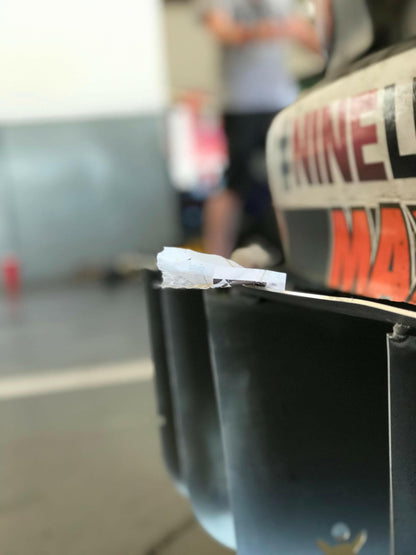 Close-up of the rear section of a vehicle, probably a car, highlighting the Miata Diffuser '90-05 NA/NB by 9livesracing with a small piece of crumpled white paper lodged in it. The background is out-of-focus, featuring people and some equipment.
