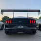 A black Mustang, fitted with the 9livesracing Big Wang Kit '14+ S550 aluminum wings to boost its down-force, is parked in a lot. The license plate reads "LOL RUN." Two other cars are partially visible on either side of the black Mustang. Trees and a building are in the background under an evening sky.