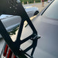 Close-up of a black Mustang's rear equipped with the 9livesracing Mustang Big Wang Kit '14+ S550, featuring large, angular aluminum wings attached to the trunk. The background shows, in soft focus, a parked car, trees, and a few people standing, highlighting a social outdoor setting that underscores high-performance engineering for maximum down-force.