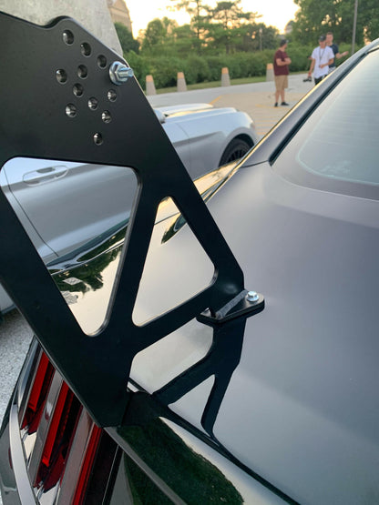 Close-up of a black Mustang's rear equipped with the 9livesracing Mustang Big Wang Kit '14+ S550, featuring large, angular aluminum wings attached to the trunk. The background shows, in soft focus, a parked car, trees, and a few people standing, highlighting a social outdoor setting that underscores high-performance engineering for maximum down-force.