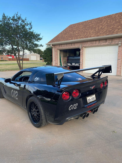 A black sports car with the number "012" painted on its sides is parked in front of a brick house with a two-car garage. The car boasts a 9livesracing Corvette Big Wang Trunk Mount Kit '97-13 C5/6 aluminum wing and Texas license plates. A sleek black pickup truck is parked inside the garage.