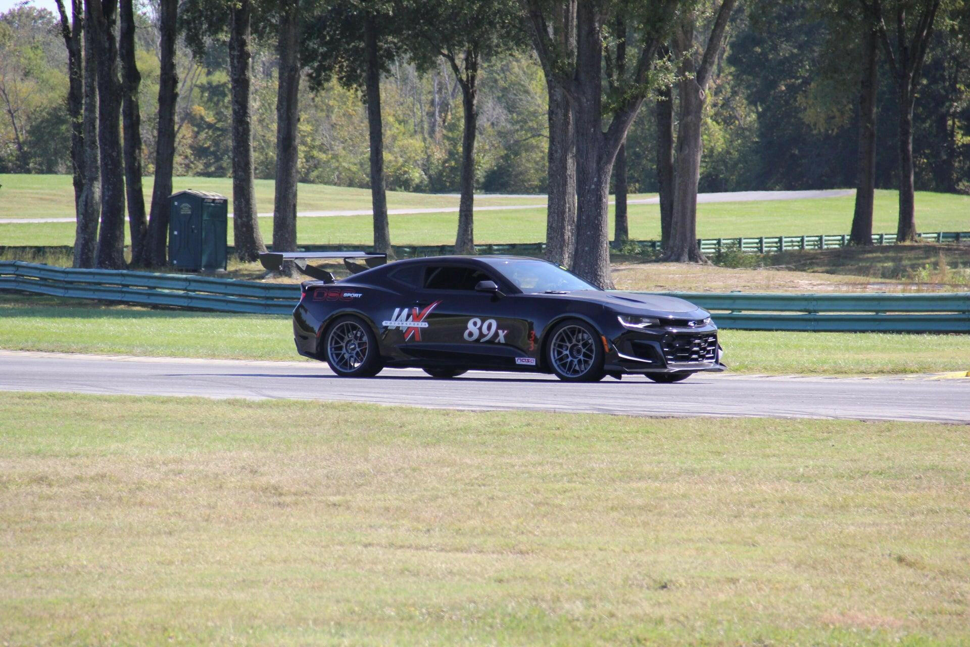 A sleek black Camaro Wang 2016+ 6th Gen from 9livesracing, adorned with the number 89X and a bold logo on its side, zooms down a paved track in a picturesque, wooded area. The car, sporting aluminum wings and a significant rear spoiler, demonstrates impressive GT3 performance. The surrounding landscape features grassy terrain with trees and fencing visible in the distance.
