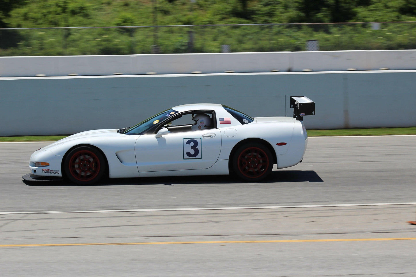 A white Corvette Rocket Nose Splitter ’97-04 C5 with the number 3 on the side speeds down a track. Featuring red wheels, a large rear spoiler for added downforce, and an American flag decal on the side, this vehiclr cuts through the air like a precise instrument from 9livesracing. A concrete barrier lines the track, and greenery is visible in the background.