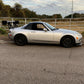 A silver sports car with a black hardtop and a Miata Big Wang Kit '06-15 NC *V1* rear wing from 9livesracing is parked on the side of the road. Situated in front of a metal gate and a grassy field, the vehicle stands out under the partly cloudy sky, embodying the spirit of 9livesracing.