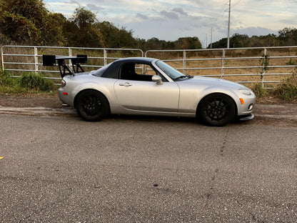 A silver sports car with a black hardtop and a Miata Big Wang Kit '06-15 NC *V1* rear wing from 9livesracing is parked on the side of the road. Situated in front of a metal gate and a grassy field, the vehicle stands out under the partly cloudy sky, embodying the spirit of 9livesracing.